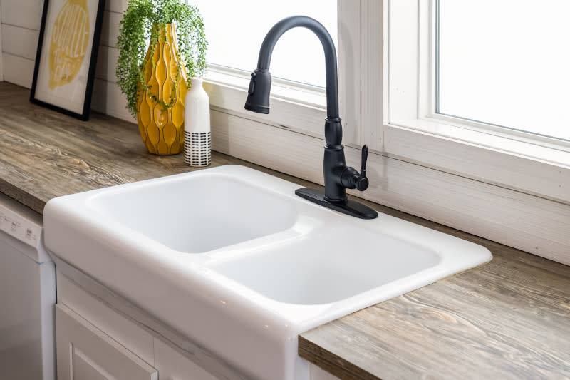 White, double basin farmhouse sink with black faucet, light wood counters with white and yellow decor and two windows behind it.