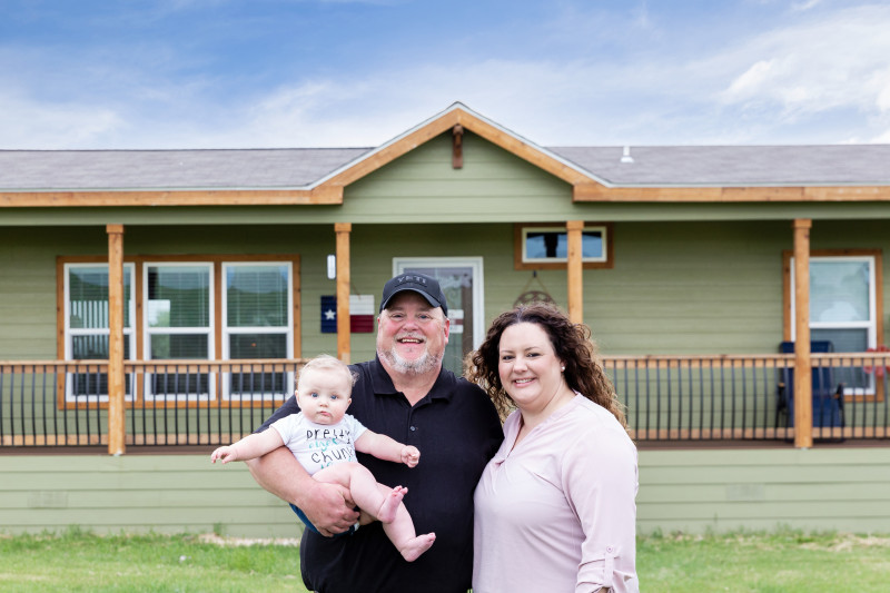 Couple stand in font of their forest green cabin home with wide front porch with man holding their new baby.
