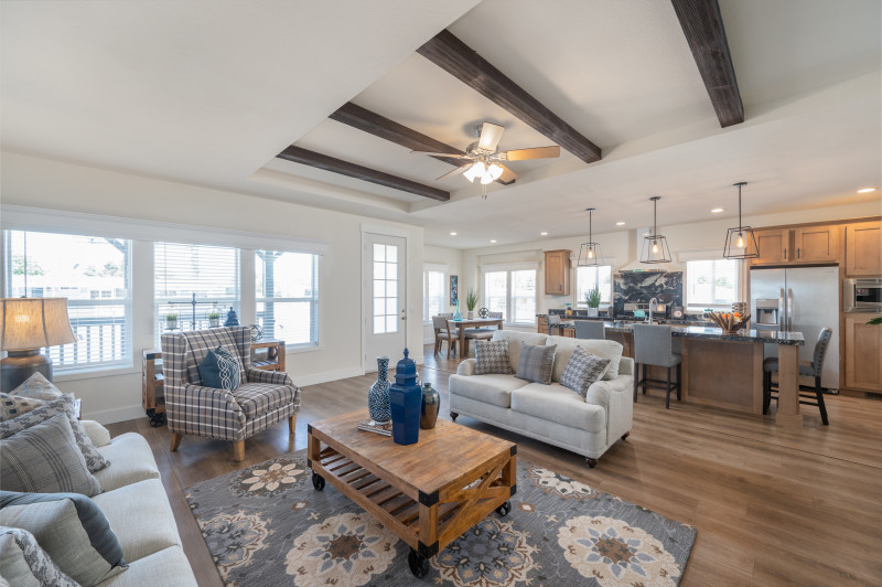 Living room of a manufactured home with gray and blue couches, chair, coffee table, rug and decor, 3 large windows and dark wood ceiling beams, with a kitchen and dining area in the background.