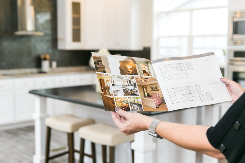 Woman wearing black shirt and silver watch holds pamphlet with home photos and layout while standing in the kitchen of a manufactured home, which is out of focus in the background.