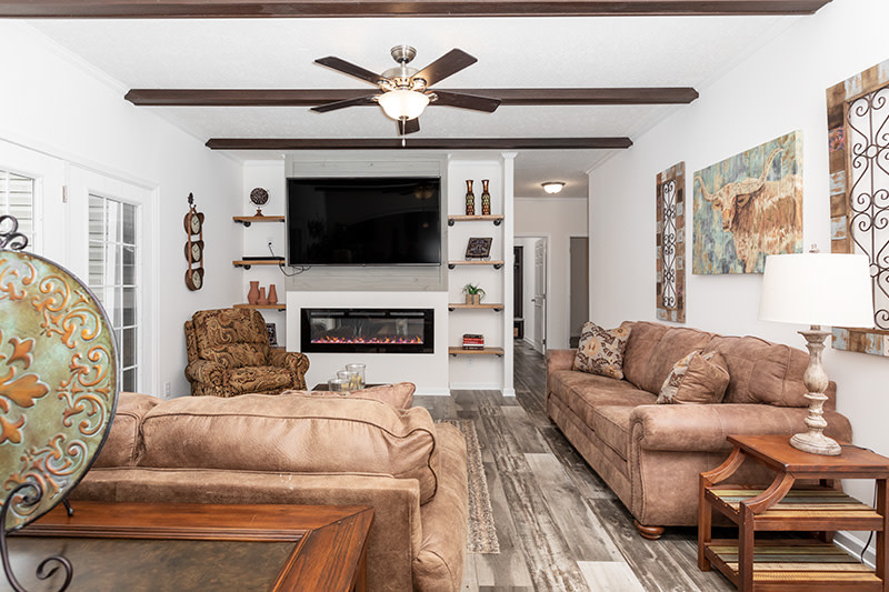 Den of a manufactured home featuring leather couches and rustic decor, with dark ceiling beams, ceiling fan and TV accent wall with electric fireplace.
