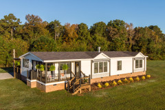 An exterior of a manufactured home with white siding, a side porch with rocking chairs and brick skirting.