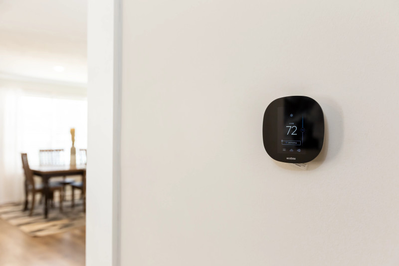 Close up of a black ecobee smart thermostat on a white wall, with a dining room table and chairs in the background