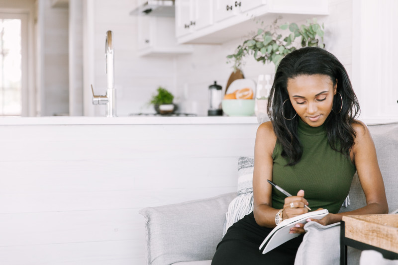 Young woman is sitting down in a chair taking notes in a notebook. She’s wearing a forest green top, and the room has natural light streaming in. 