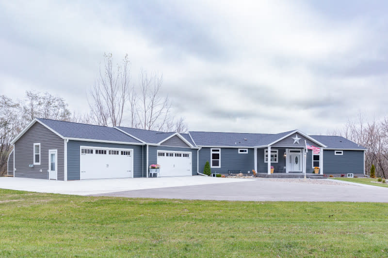 Manufactured home against cloudy sky, with blue-gray siding, white trim and doors and 2 attached 1-car garages.