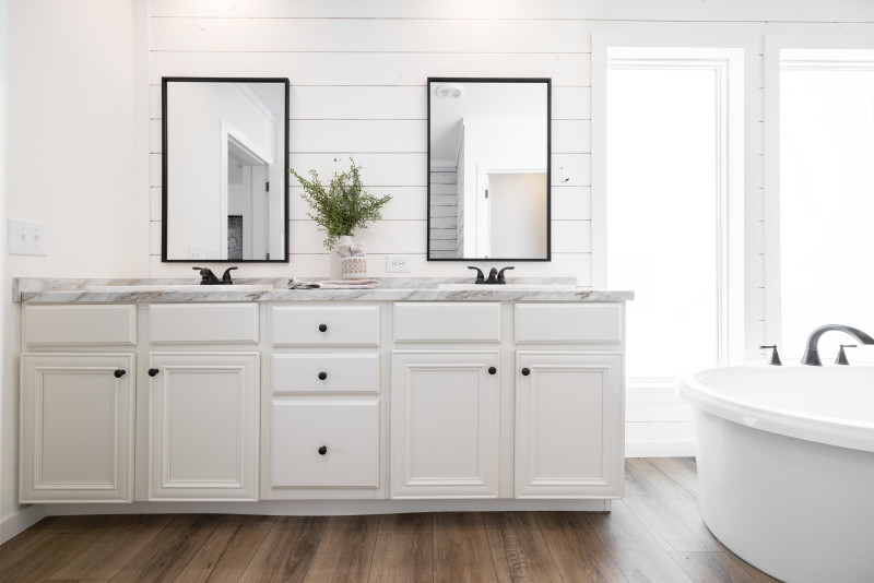  A bright white, primary bathroom with double vanity that has black framed mirrors hanging over it. Larger windows are to the right with part of a freestanding tub in view.