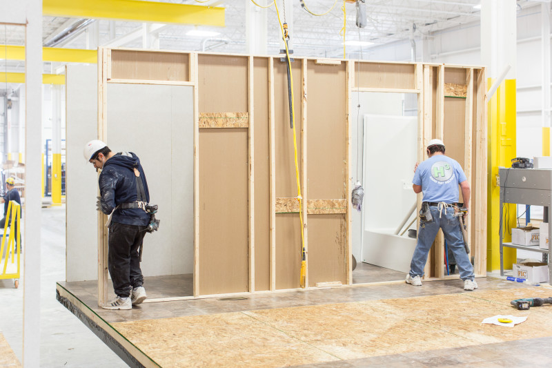 Two men install interior wall of a manufactured home inside of a home building facility.