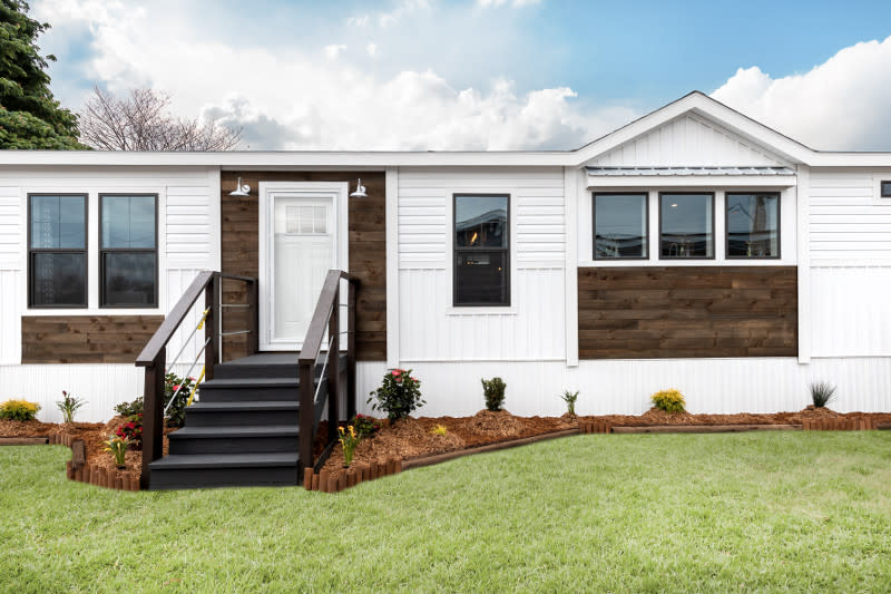 Exterior of a manufactured home with white siding, dark wood accents, white front door and black steps leading to the grass of the yard.