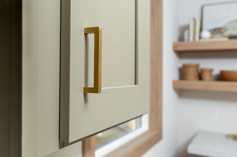 Closeup of a sage green kitchen cabinet in a manufactured home with brass handle and light wood trim and shelves in the background.