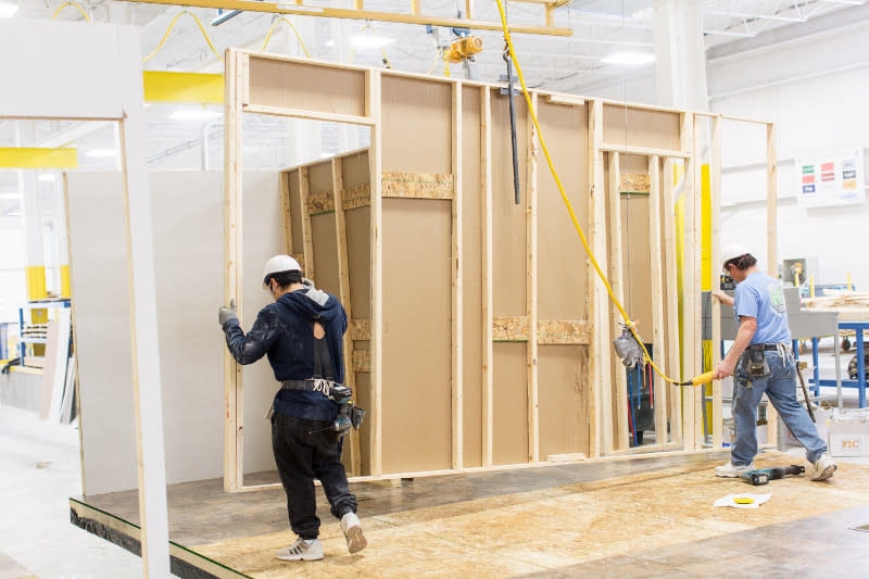 Two team members work to assemble wall of manufactured home inside a home building facility.