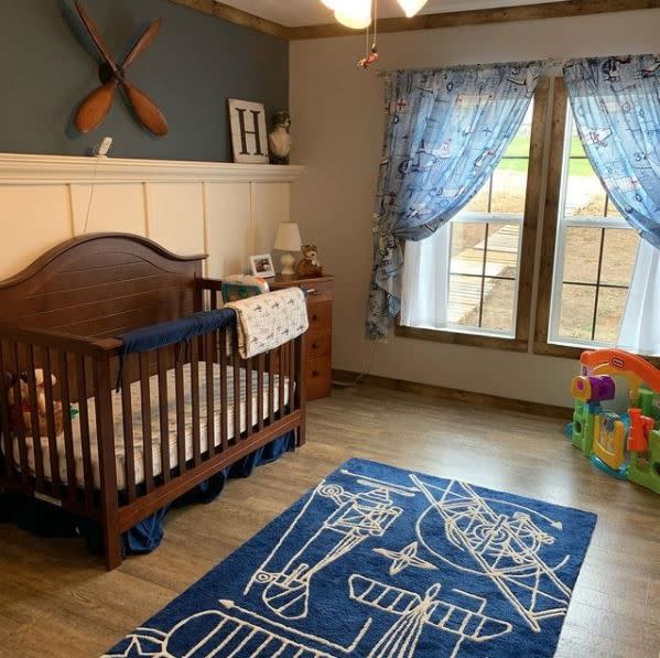 Nursery with dark wood crib and table, white wainscotting, dark green wall, blue rug and light blue curtains.