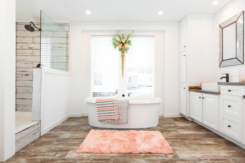 A bathroom with sink and white cabinets on one side, a large oval bathtub with a window behind it in the middle and a half-wall glass shower on the other side.