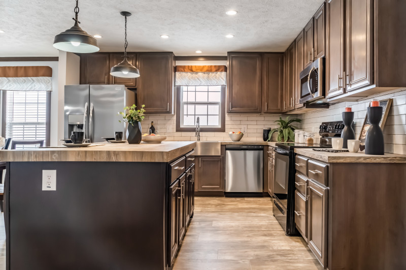 The kitchen in this manufactured home has dark wooden cabinets and an island. There are also some stainless-steel appliances and white subway tiles as the backsplash.