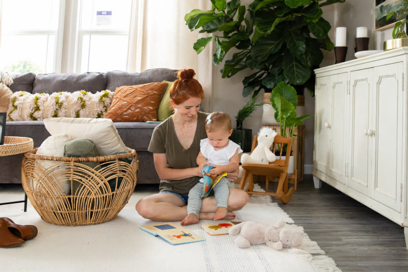 Woman sits with young child holding a book on a white rug on the floor of a manufactured home living room with toys, basket holding pillows, gray couch with pillows, plants and white sideboard behind them.