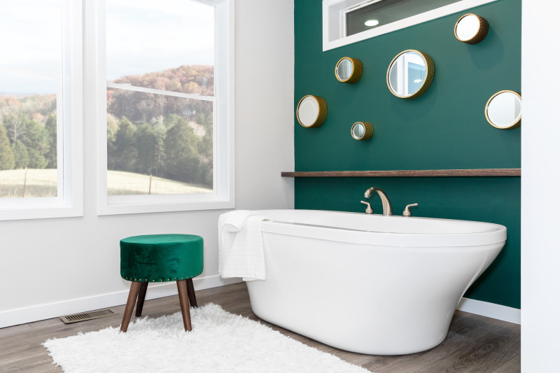 Bathroom of a Clayton Built home with soaker tub, green velvet stool, two large windows and green accent wall with mirrors on it.