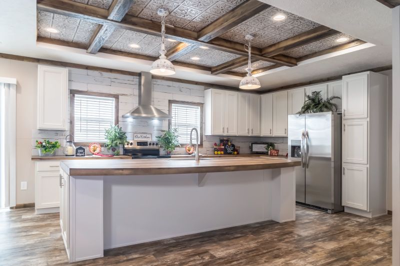 A manufactured home kitchen with a large white island, white farmhouse cabinetry and a tin and wooden beamed ceiling.