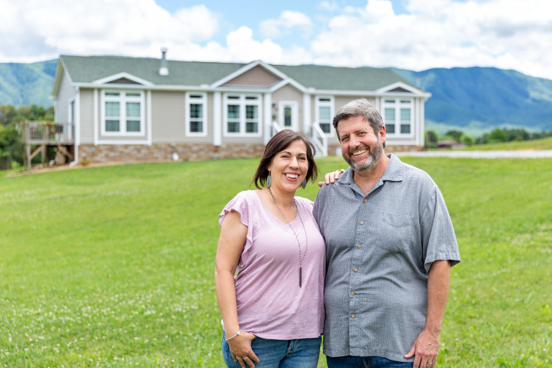 Man and woman stand together smiling with modular home exterior and East Tennessee mountains in the background. 