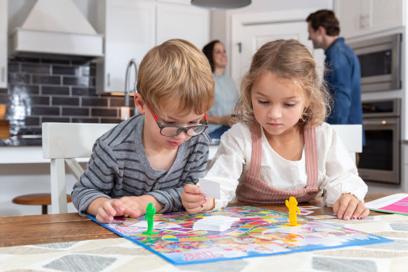 A young boy and girl playing a board game while their parents are in the kitchen.