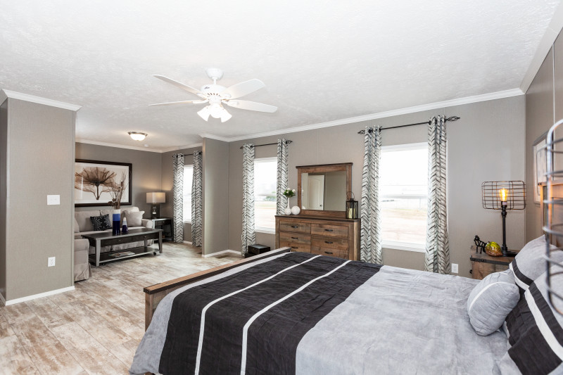 Primary bedroom suite of a manufactured home with light wood floors, beige walls, gray, black and wood decor and a nook with a couch and coffee table.