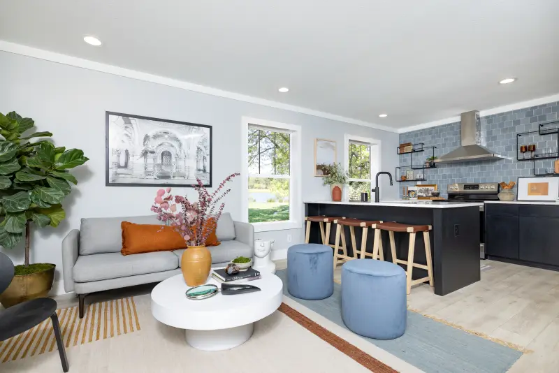 A Clayton eBuilt home's living room with a modern couch and round white coffee table leading to a kitchen with a breakfast bar, dark wood cabinets and a blue subway tile backsplash.