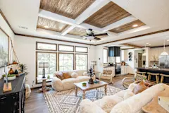 A living room with fluffy beige couches, a tray ceiling with white ceiling beams, three large windows and a rug with a neutral geometric pattern; a spacious kitchen is in the background to the right.