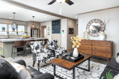 View of the Bluebonnet Breeze. An open floor plan is displayed with both the living room and kitchen in view. Kitchen has a large island and chalkboard. Living area features faux-cow print chairs, wooden coffee table and dresser. 