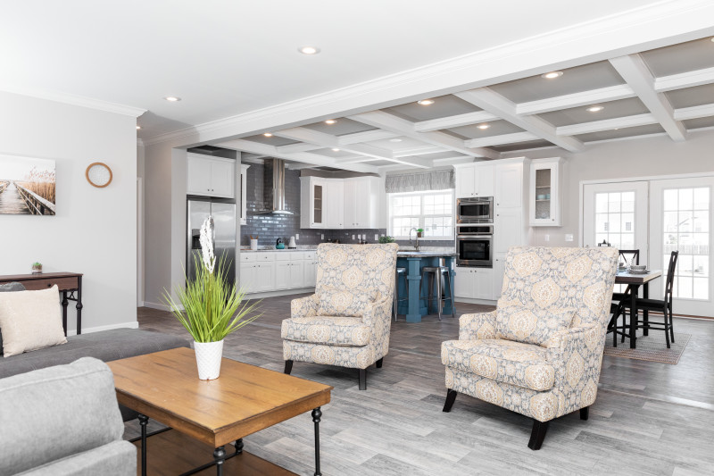 Living room of a manufactured home with light gray floor, 2 chairs and coffee table, with a white kitchen and dining area behind and a coffered ceiling.