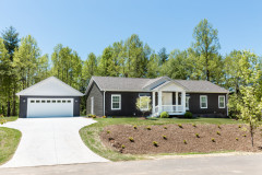 Manufactured home with dark brown siding, white trim and porch and matching detached garage with driveway and lawn.