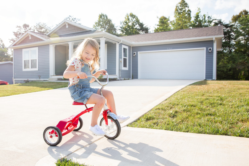 Child riding a red tricycle on the sidewalk outside a blue manufactured house.