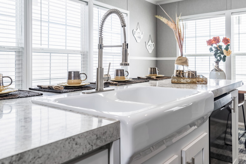 View is of a while sink with silver, gooseneck, detachable nozzle on a white marble-style countertop island. There are place settings with brown mugs, plates, utensils, and placemats.