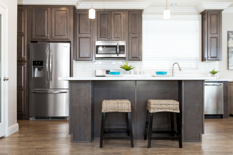 Kitchen of a manufactured home with dark wood cabinets, lighter wood floor, stainless streel appliances, subway tile backsplash and island with 2 stools.
