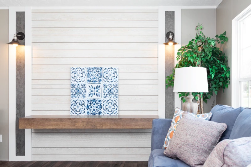 A white, ship-lap accent wall with shelf is in view with a blue art piece. There is a decorative tree, couch, and light in the background. 