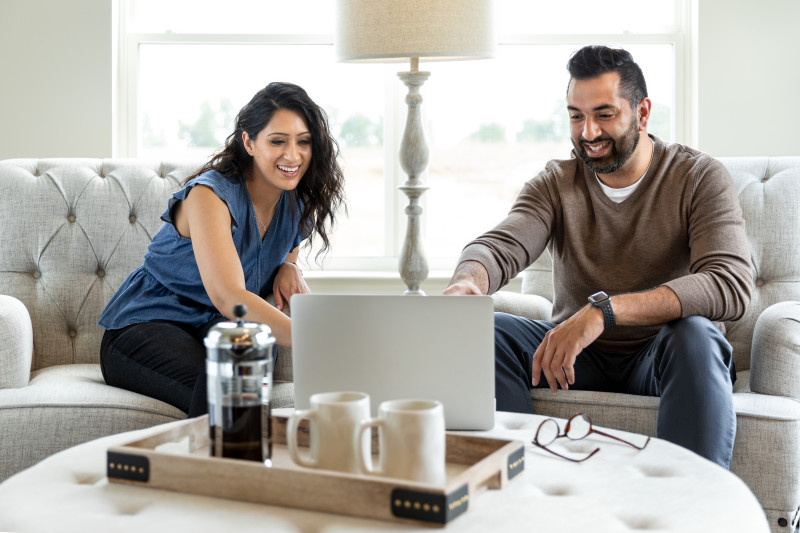 Woman and man sit on beige chairs in front of window, pointing at a laptop on a beige ottoman with a tray with coffee mugs and French press in front of it.