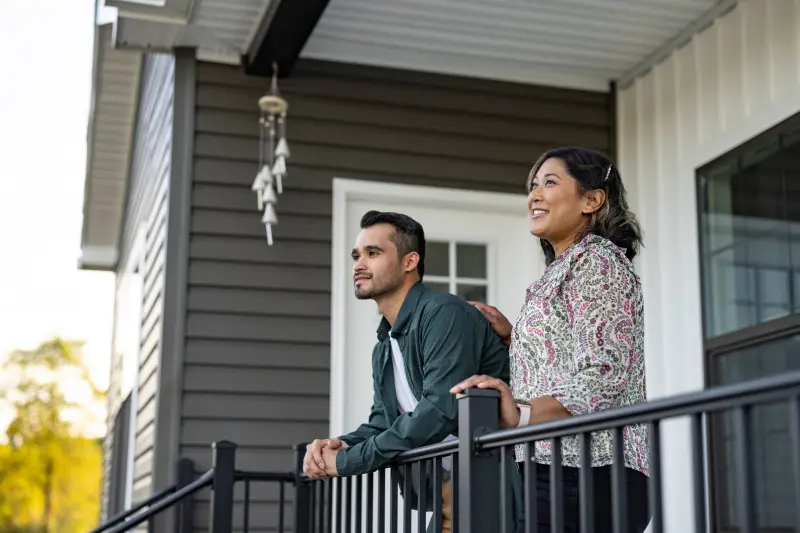 Man and a woman stand on the porch of a manufactured home with dark gray siding and white trim