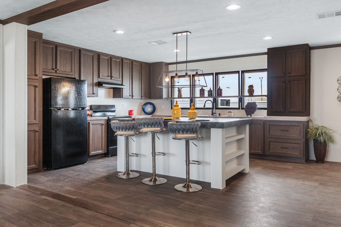 The kitchen of a Clayton manufactured home with dark wood cabinets and floors, and island with seating and a chandelier, and a row of four windows.