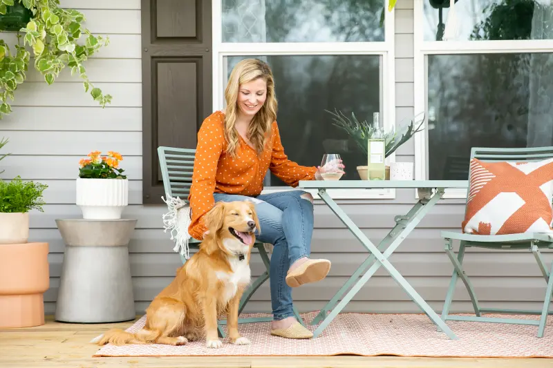 A woman on porch pets her dog and holds a wine glass.