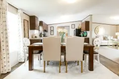 Dining area in a manufactured home with a wooden table and white chairs. A kitchen and living area are in the background. 