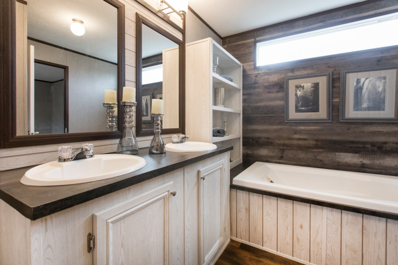Primary bathroom of a single wide manufactured home with wood accent wall, storage shelves, tub with shiplap accent, shiplap accent wall and vanity with 2 sinks and 2 mirrors over it.
