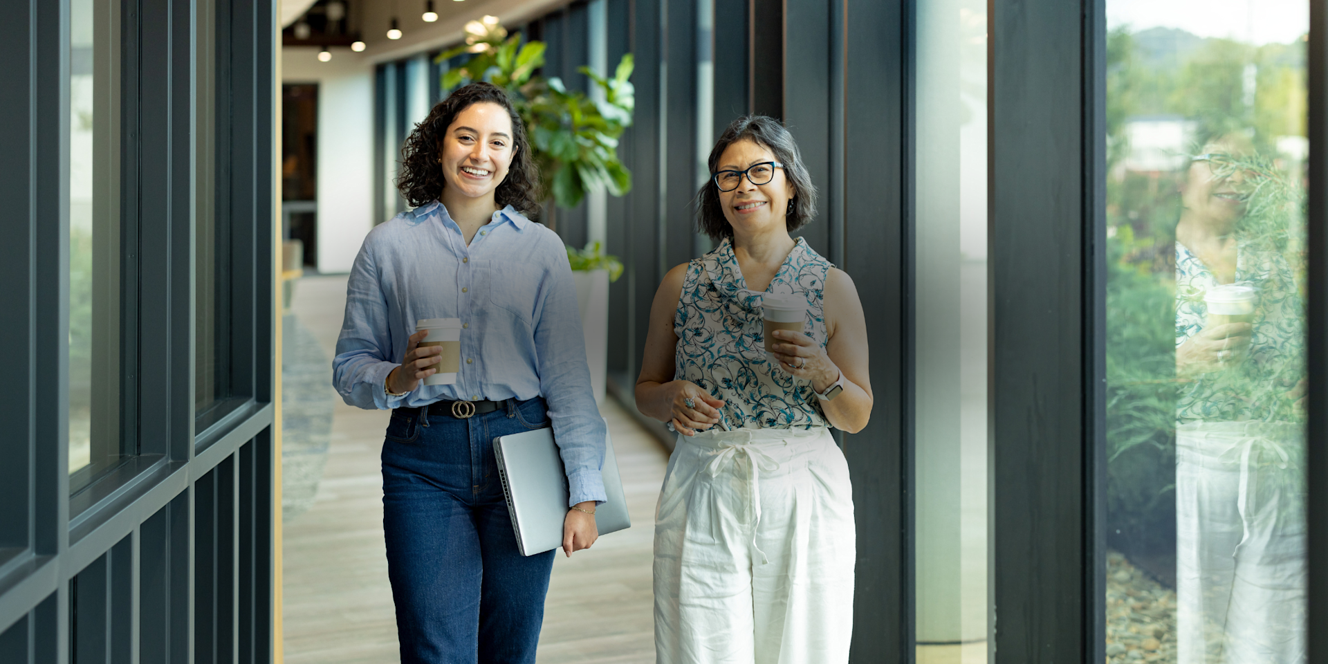 Two women walk in an office hallway beside a wall of windows and carry paper coffee cups.