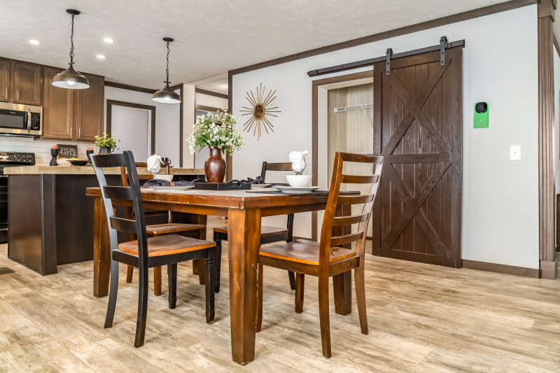 An angled view of the dining area in a manufactured home shows a table and chairs with a pantry in the background. The kitchen is in partial view to the left with an island and light fixtures hanging down over it.