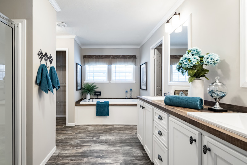 View of a bathroom with a double vanity, walk-in shower, and a soaker tub with windows situated above it. There’s a linen closet in the distance. Bathroom is creme colored with teal towels to accent.