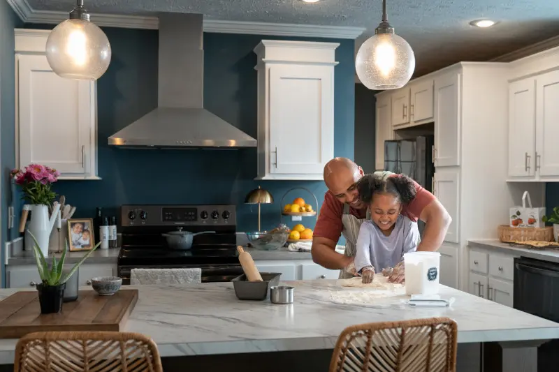 Man and girl make cookie dough at the kitchen island of a manufactured home, with a blue wall and white cabinets.
