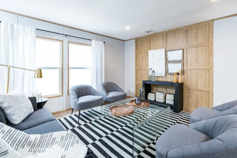 Interior of the Morocco home model with large windows and sheer white curtains. Home has wooden accent wall and is decorated with a black and white striped carpet, clear table and four gray chairs. 