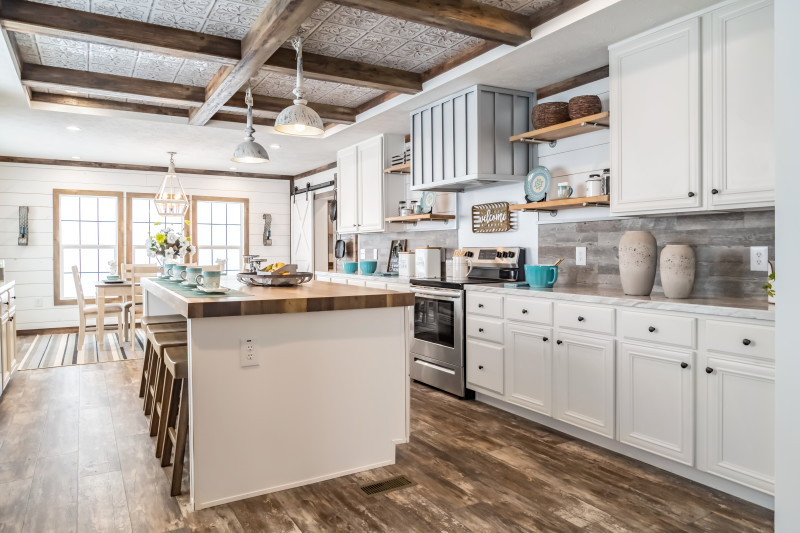 A farmhouse kitchen with white cabinets, a large island with butcher block-style counters and a coffered ceiling, with a bright dining room in the background.
