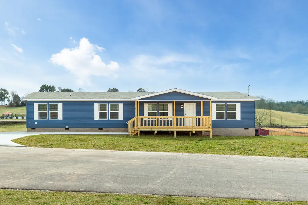 A blue manufactured home exterior with white shutters and door, a wooden front porch and stone skirting sits on a green yard with off a driveway, a rural scene of trees and gentle green hills in the background, under a bright blue sky.