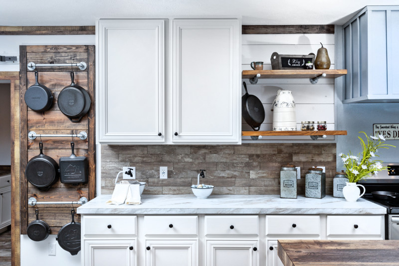 Farmhouse kitchen with wood grain hanging rack holding cast iron skillets, white cabinets, marble style countertops, and stone backsplash.