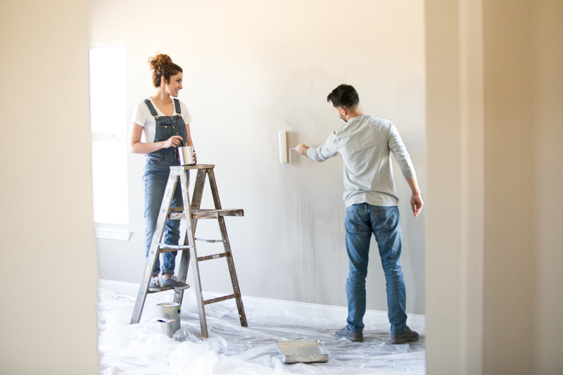 A couple paints an empty room with plastic on the floor; the one on the right wear a grey shirt and jeans while using a pain roller on the wall while the other stands on a ladder in overalls, stirring paint in a can.