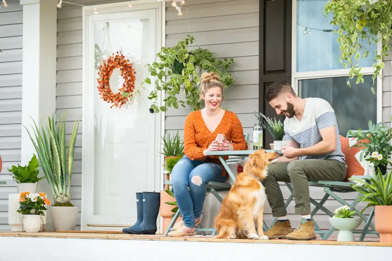 Man and woman on patio furniture with a dog on the front porch of a tan manufactured home, surrounded with plants.
