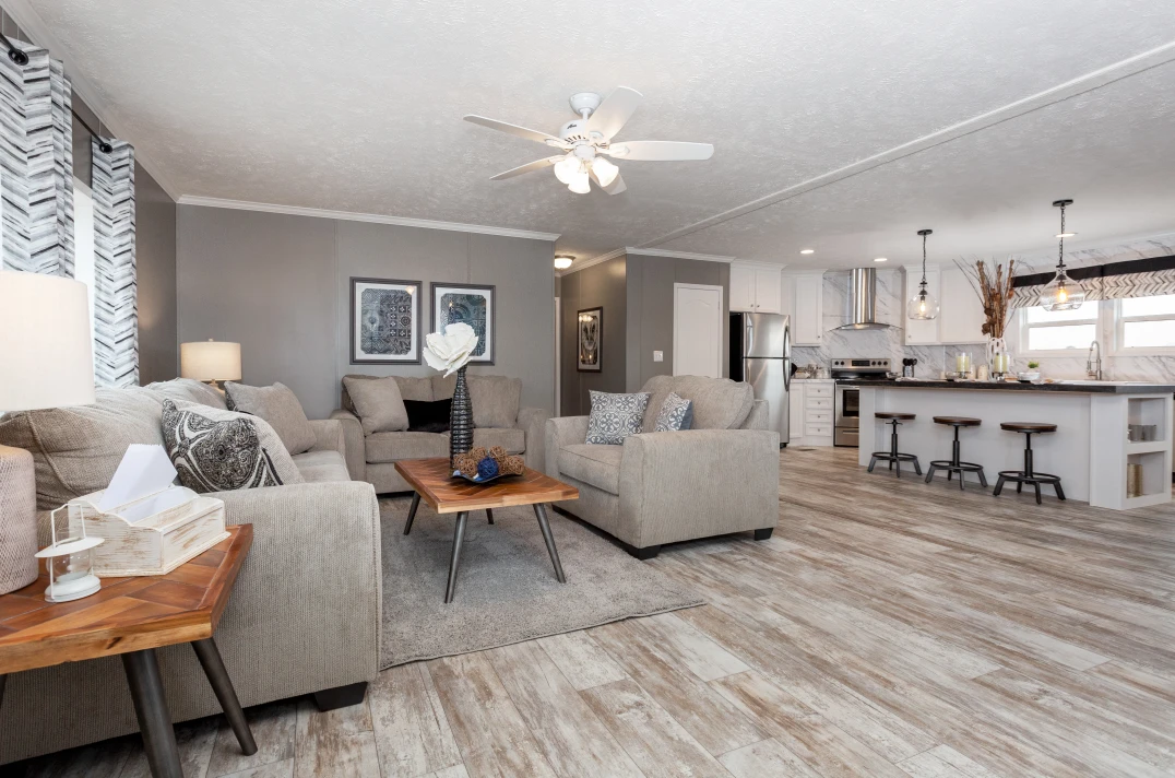 A living room with beige sofas around a wood coffee table is open to the kitchen featuring a large white kitchen island and stainless steel appliances.