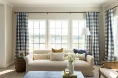 Three large windows are framed by blue checkered curtains and are behind a cream couch in a living room.